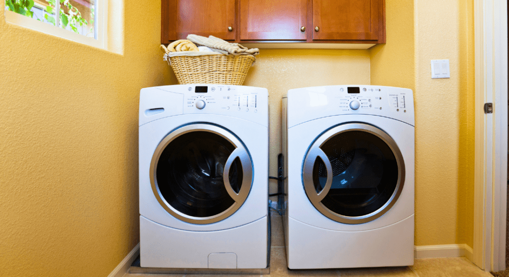 A laundry room with a front-loading washer and dryer side by side, a basket with folded towels on top, and wooden cabinets above.