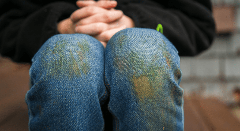 Close-up of a seated person wearing jeans with visible dirt stains on the knees, hands clasped together resting on their lap. Grass stains