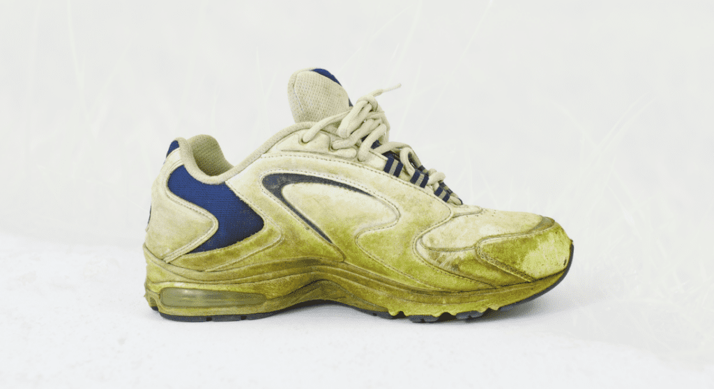 A worn-out, dirty athletic shoe with yellow and green discoloration and signs of wear, set against a light background. Grass Stains