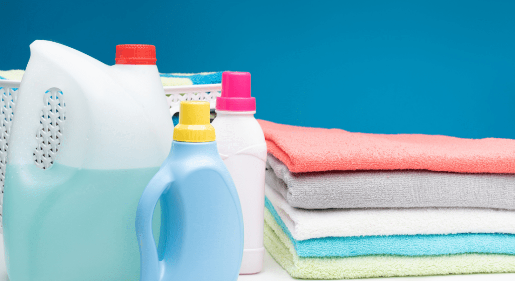 Various detergent bottles are placed on a surface next to neatly folded colorful towels with a laundry basket in the background. Laundry Myths