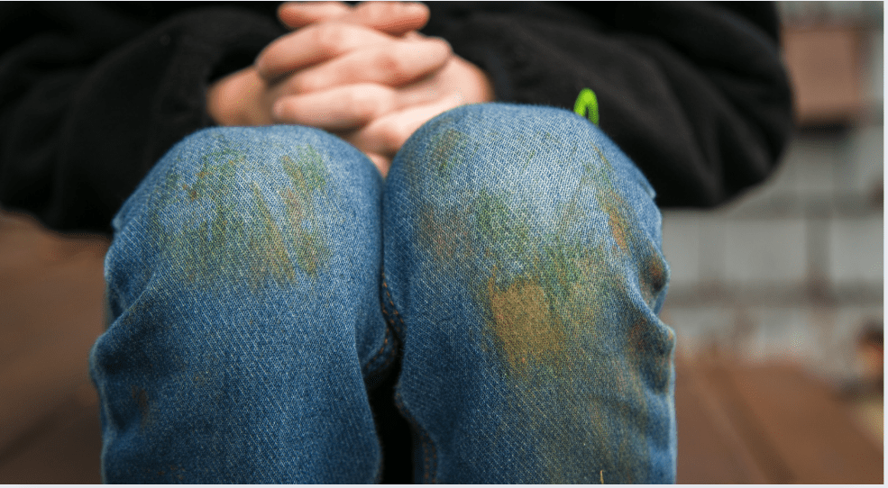 Close-up of a person’s hands clasped above their knees, with blue jeans showing visible grass stains and dirt marks.