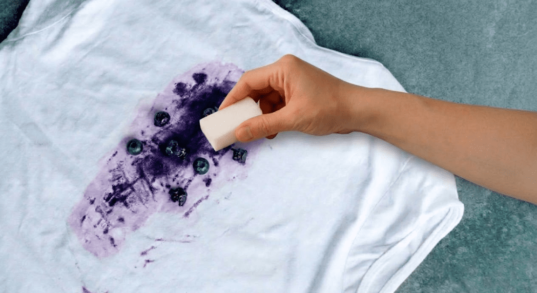 A hand uses a white eraser to clean a large purple berry stain on a white t-shirt lying on a flat surface.