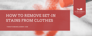 Close-up of a red stain on white fabric with a red cleaning brush. Text reads, "How to remove set-in stains from clothes. thedutchmanslaundry.com.