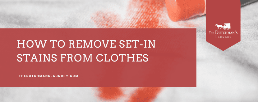 Close-up of a red stain on white fabric with a red cleaning brush. Text reads, "How to remove set-in stains from clothes. thedutchmanslaundry.com.