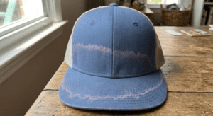 A blue and beige baseball cap with a faded pink line across the front sits on a wooden table near a window.