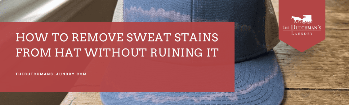 A blue and gray hat with visible white sweat stains on the brim and front, placed on a wooden surface next to a banner with stain removal advice.